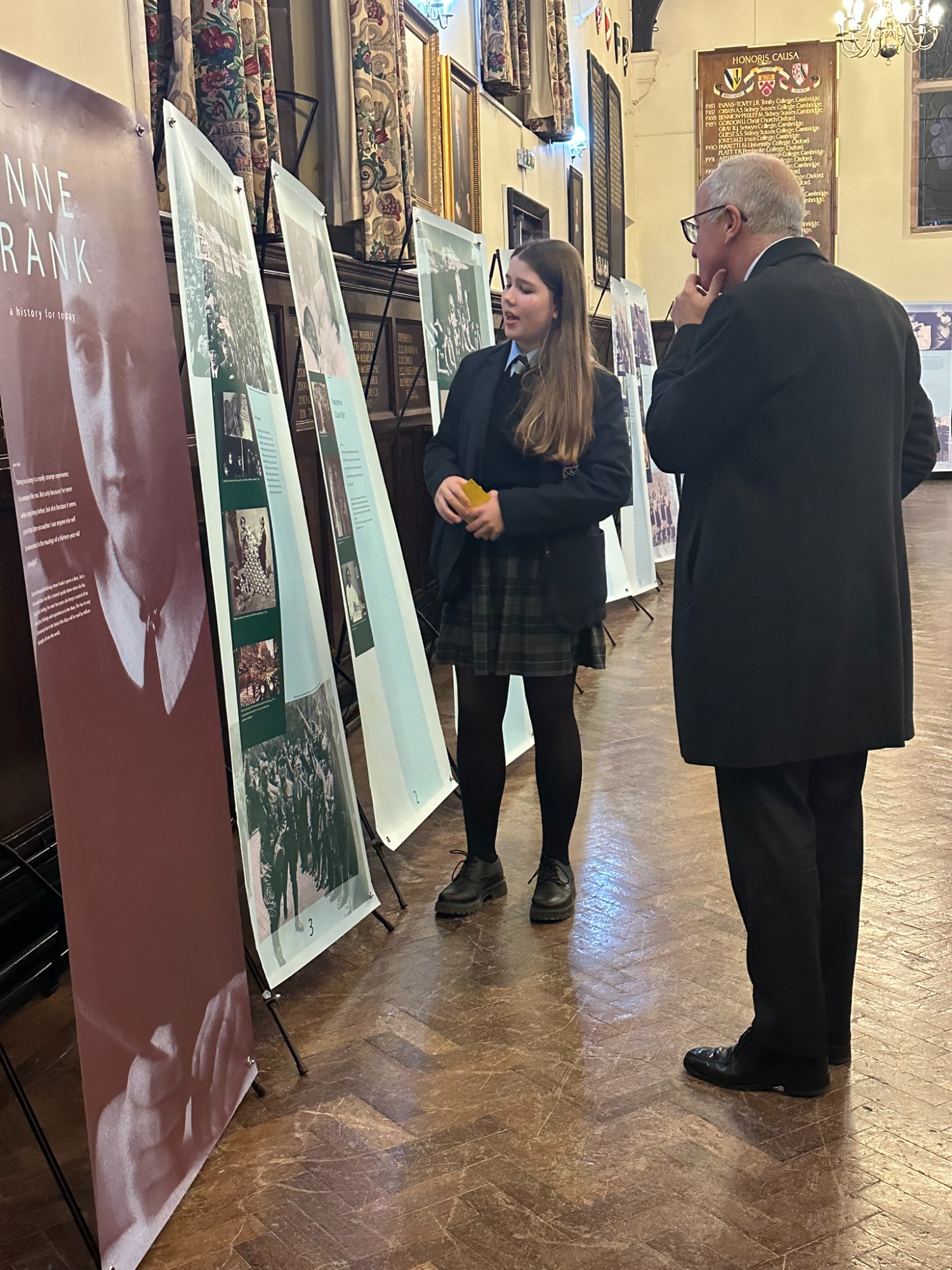 Female student stands with a male visitors infront of exhibition stands in the grand Great Hall, talking him through the exhibition