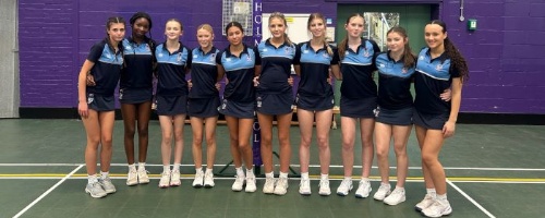 10 girls in sports kit line up for team photo in a sports hall in front of a netball hoop