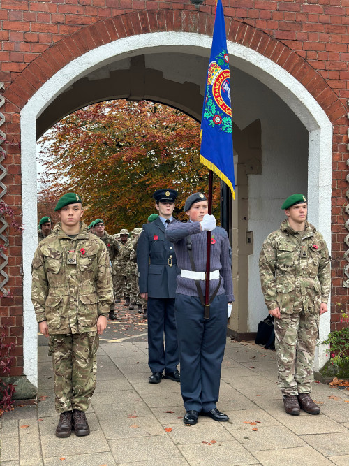 student cadets in army and air force uniforms bearing a flag, standing beneath a brick archway with student cadets dressed in military fatigues lined up behind them